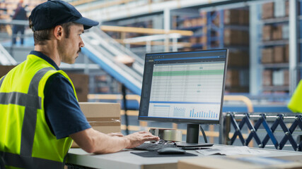 Male Stocking Associate Checking Inventory On Desktop Computer In Warehouse Facility With Automated...