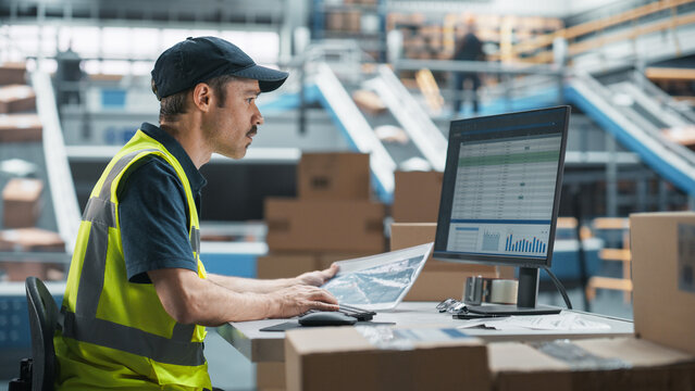 Male Stocking Associate Checking Inventory On Desktop Computer In Warehouse Facility With Automated Conveyor Belt. Sorting Center Employees Carrying Boxes to Package Products, Deliver to Clients.