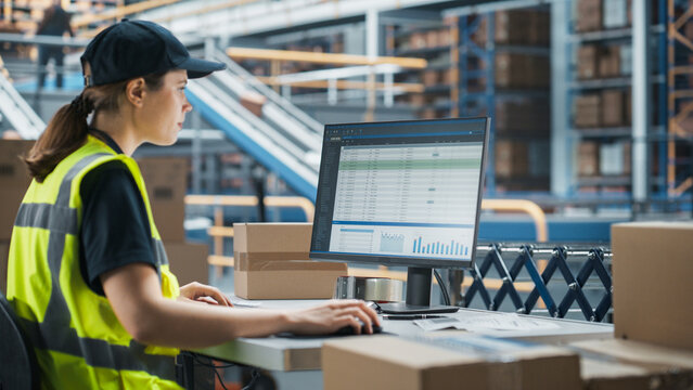 Caucasian Female Stocking Associate Using Desktop Computer To Check Inventory In Distribution Facility. Multiethnic Colleague Loading Cardboard Boxes With Online Orders On Automated Conveyor Belt - Powered by Adobe