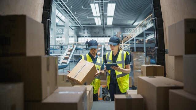 Caucasian Female Stocking Associate Using Tablet Computer To Check Inventory In Logistics Facility With Conveyor Belt. Male Warehouse Worker Loading Boxes In Van For Delivering Orders To Clients.
