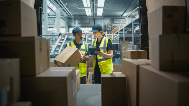 Caucasian Female Supervisor Using Tablet Computer To Check Inventory In Modern Logistics Facility With Conveyor Belt. Male Warehouse Worker Loading Boxes In Van For Delivering Orders To Clients.