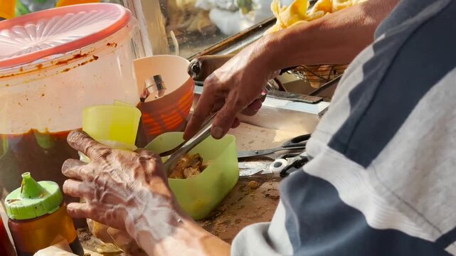 Street food seller preparing Batagor, a popular Indonesian fried dumpling with peanut sauce, using scissors and tongs at a traditional food stall in daylight