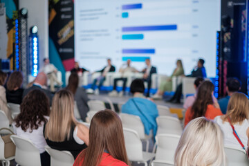 Professionals attending a business conference, seated in an audience with presentations on stage.
