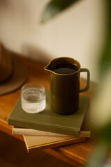 Close-up of a ceramic jug filled with water and a glass of water on stacked books in warm natural light. Cozy and minimal home setting with soft shadows.	