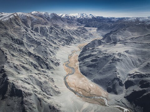 Aerial view of a winding, sandy river snakes through a rugged valley, framed by stark, snow-dusted mountain peaks under a vast blue sky, Turgen, Uvs, Mongolia.