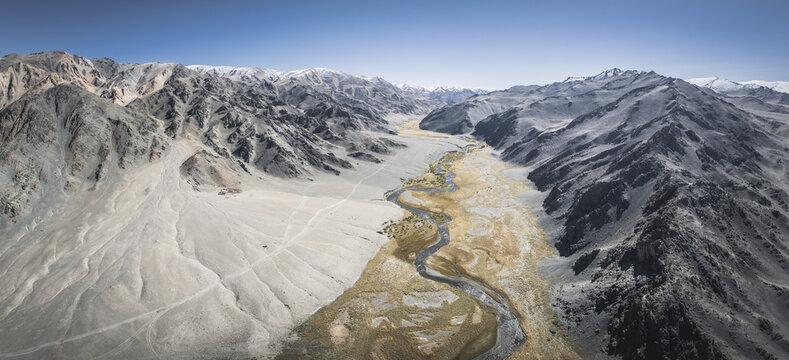 Aerial view of a meandering river carves through a vast, arid valley, framed by stark, rocky mountains under a clear sky, Turgen, Uvs, Mongolia.