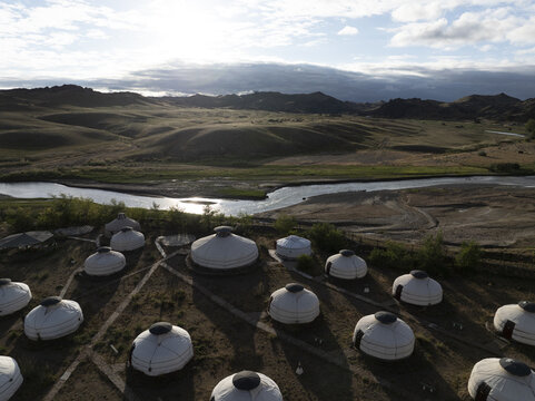 Aerial view of a cluster of traditional yurts nestled near a winding river, against a backdrop of rolling hills under a soft, hazy sky, Turgen, Uvs, Mongolia.
