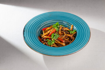 Korean style stir fried udon noodles with beef, vegetables, cilantro and sesame served in a blue rustic plate on white tablecloth background