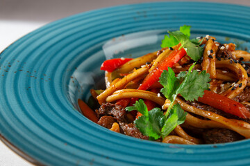 Korean style stir fried udon noodles with beef, vegetables, cilantro and sesame served in a blue rustic plate on white tablecloth background