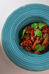 Traditional Korean japchae noodles with beef, bell pepper, carrot, sesame seeds and cilantro served in a rustic blue ceramic plate on white background