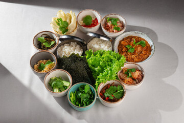 Top view of a traditional Korean meal setup with assorted banchan side dishes, steamed rice, fresh lettuce, seaweed sheets and spicy stew, served on a light background
