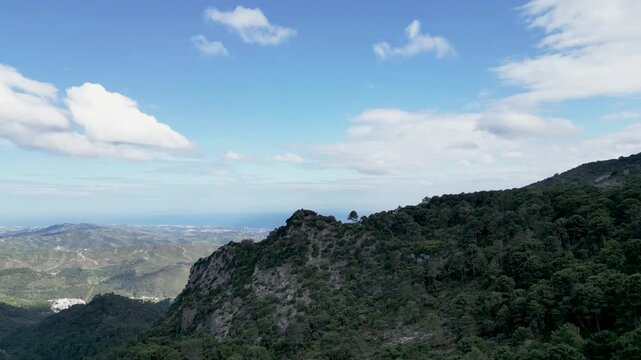 Aerial view of rugged mountain ridges covered in lush green vegetation under a blue sky spotted with white clouds, Ojen, Marbella, Spain.