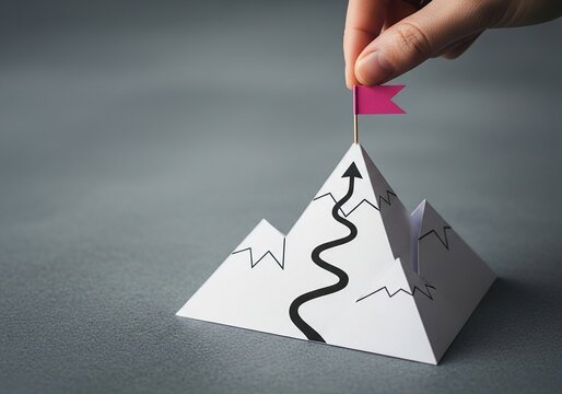 Hand places pink flag on paper mountain peak with winding path to success