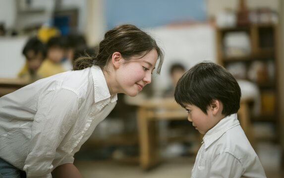 A young East Asian teacher looks at a child with a gentle smile in an art classroom setting. A tender moment of connection and care