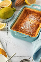 Baked lemon pudding with lemon zest and juice garnished with powdered sugar in blue baking dish on white tile background