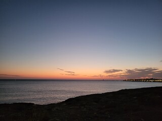 Tranquil coastal sunset over calm sea with silhouette of shoreline and lighthouse under vibrant orange and blue evening sky