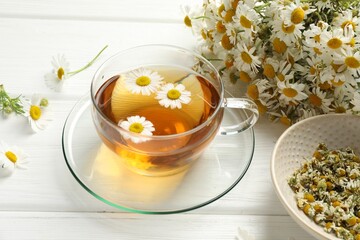 Aromatic tea in glass cup and chamomile flowers on white wooden table, closeup