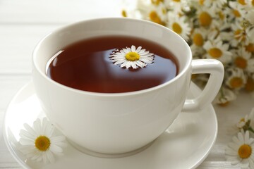 Aromatic tea in cup and chamomile flowers on white table, closeup