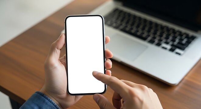Person holding a smartphone with a blank white screen, laptop nearby