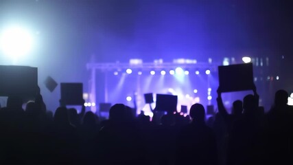 Crowd at a nighttime concert holding signs. Blurred silhouettes of people in the audience, illuminated by vibrant purple and blue stage lights