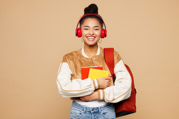 Young smiling happy smart girl student wear casual clothes backpack bag hold books listen to music in headphones isolated on plain pastel light beige background High school university college concept