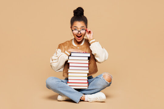 Full length young fun surprised girl student wear casual clothes glasses backpack bag sitting hold stack of many paper books isolated on plain beige background. High school university college concept.
