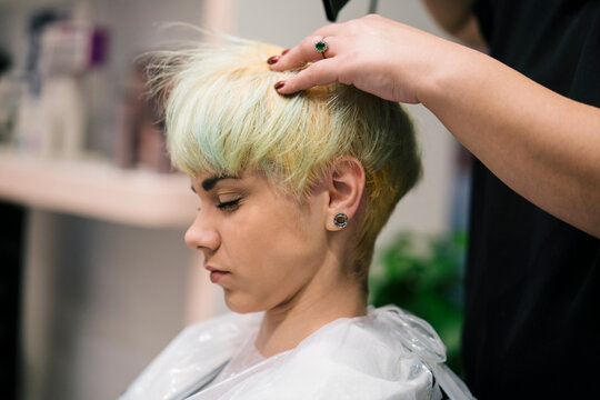 A young woman with short, blonde and green hair sits in a salon chair. A hairdresser's hand is seen styling or drying her hair. The client wears a protective cape during the process.