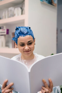 A young woman with blue hair dye smiles while reading a book in a salon. She wears a protective cape during her hair coloring process.