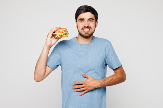 Young man in blue t-shirt casual clothes hold eat burger put hands on abdomen suffer from stomach ache isolated on plain solid white background Proper nutrition healthy food unhealthy choice concept - Powered by Adobe