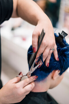 A hairdresser with red nail polish is cutting the vibrant blue hair of a client in a salon. The stylist uses scissors and a comb to trim the strands.