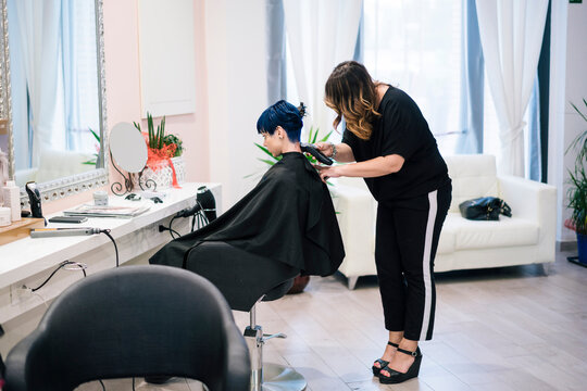 A female hairdresser trims a client's blue hair with an electric clipper in a bright, modern hair salon. The client sits calmly in the salon chair while the stylist performs the precise haircut.