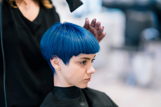 A young woman with blue hair is getting her short haircut styled by a hairdresser in a salon. The stylist's hand is near her head, shaping the vibrant blue hair.