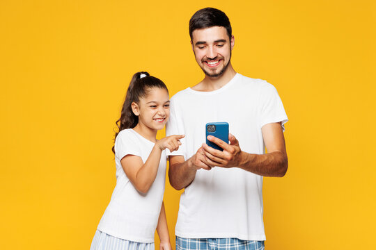 Young parent man with child girl wear casual blue pyjamas clothes. Daddy little kid daughter hold use point on mobile cell phone isolated on plain yellow background. Father's Day Love family concept. - Powered by Adobe
