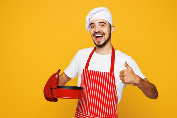 Young housewife housekeeper chef cook baker man wear red apron toque hat hold metal baking form for pie show thumb up isolated on plain yellow orange background studio portrait. Cooking food concept.