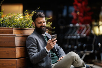 Portrait of happy classy businessman sitting on bench on a city street with mobile phone and texting messages.