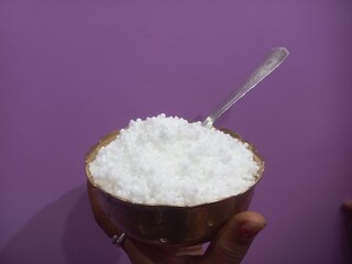 This image shows a metal bowl filled with white sago pearls (sabudana) placed inside a larger brass plate.A spoon rests inside the bowl, scooping some of the sago pearls.