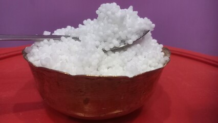 This image shows a metal bowl filled with white sago pearls (sabudana) placed inside a larger brass plate.A spoon rests inside the bowl, scooping some of the sago pearls.