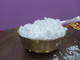 This image shows a metal bowl filled with white sago pearls (sabudana) placed inside a larger brass plate.A spoon rests inside the bowl, scooping some of the sago pearls.