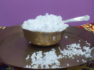 This image shows a metal bowl filled with white sago pearls (sabudana) placed inside a larger brass plate.A spoon rests inside the bowl, scooping some of the sago pearls.