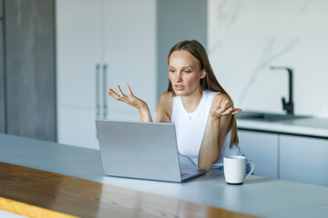 Angry shocked young woman looking at laptop screen, sitting at table in modern kitchen at home