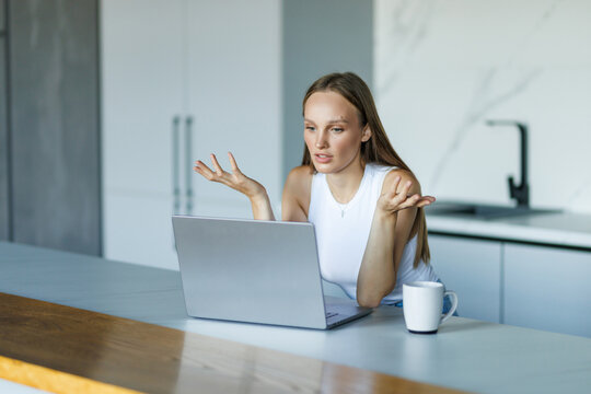 Angry shocked young woman looking at laptop screen, sitting at table in modern kitchen at home