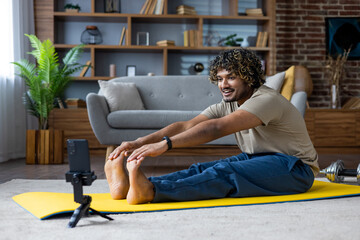 Young man performing stretches on a yellow yoga mat indoors, accompanied by a mobile phone for...