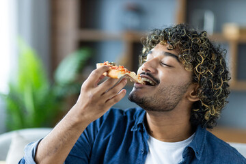 A young man with curly hair enjoys a slice of pizza, savoring the taste in a comfortable home...