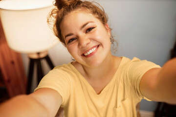 Upper view of caucasian smiling plus size millennial woman vlogger with hair bun spending leisure time at home taking selfie, recording new content to vlog, stretching arms to camera