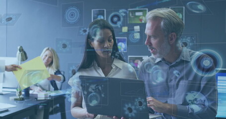 Analyzing two colleagues in business attire pointing at laptop screen in office, with data overlays