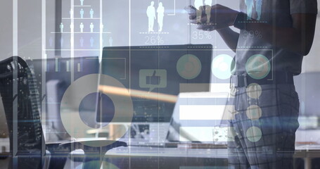 Holding smartphone worker standing beside desk in office with laptop-monitor and graphs, copy space