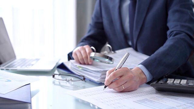 Businessman using a calculator and magnifying glass while analyzing financial documents at desk in modern office. Audit and taxes in business