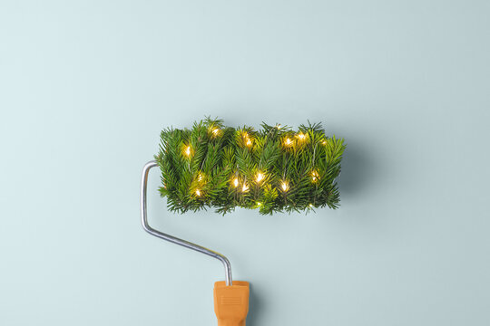 A paint roller wrapped in spruce with warm fairy lights promises a festive home makeover on a soft blue background this winter. - Powered by Adobe
