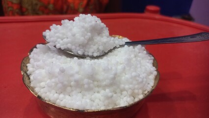 
A brass bowl filled with white sago pearls (sabudana) and a spoon rests on a red perforated surface, showing the pearls’ small, round texture clearly.
A brass bowl filled with white sago pearls (sa

