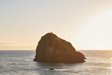 Golden hour illuminates a dramatic sea stack rising from the deep blue ocean against a soft, bright sky.